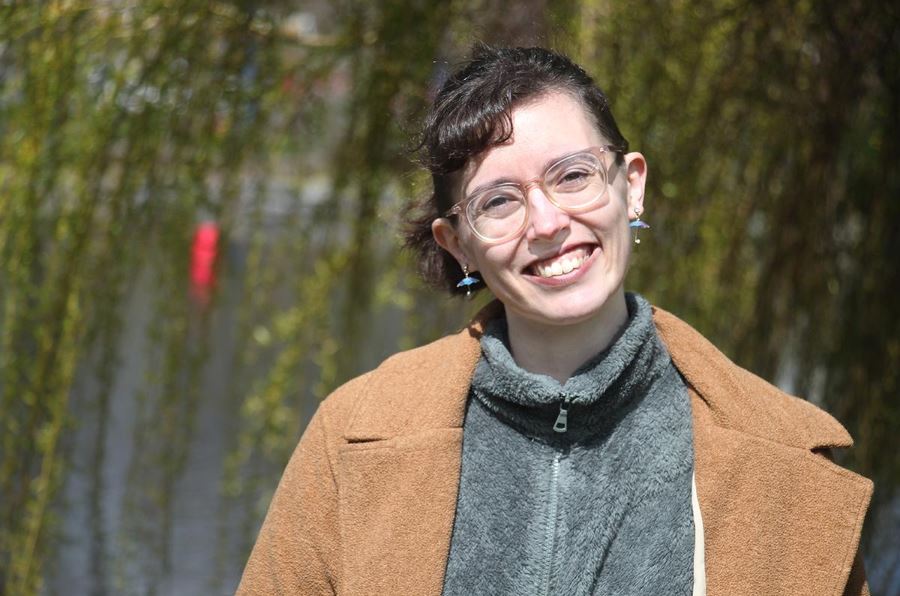 a photo of washington state poet laureate arianne true smiling at the camera. arianne wears glasses and is standing outsdie with greenery in thebackground. they wear gray fleece and a camel colored coat.